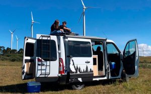 Casal sentado em cima do seu motorhome estacionado com as portas abertas, pois é sua casa sobre rodas.