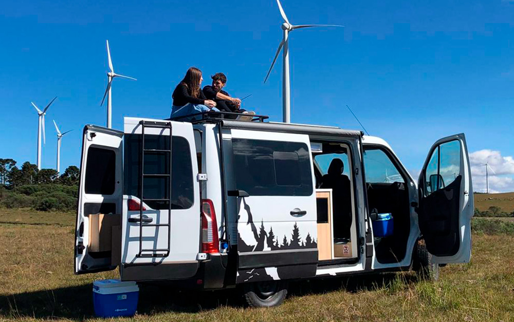 Casal sentado em cima do seu motorhome estacionado com as portas abertas, pois é sua casa sobre rodas.