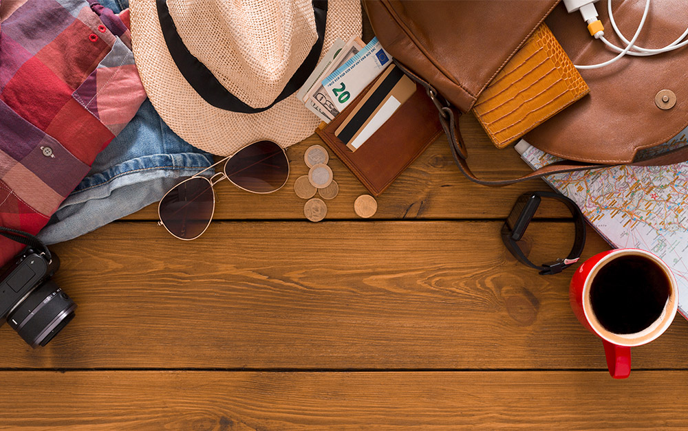 Fundo amadeirado com diversos itens exemplificando o que levar em uma viagem de motorhome, da esquerda para a direita: câmera fotográfica, roupas (xadrez e jeans), chapéu, óculos de sol, moedas, carteira com notas de dinheiro, bolsa, carregador de celular, relógio de pulso digital, mapa e xícara com café preto.
